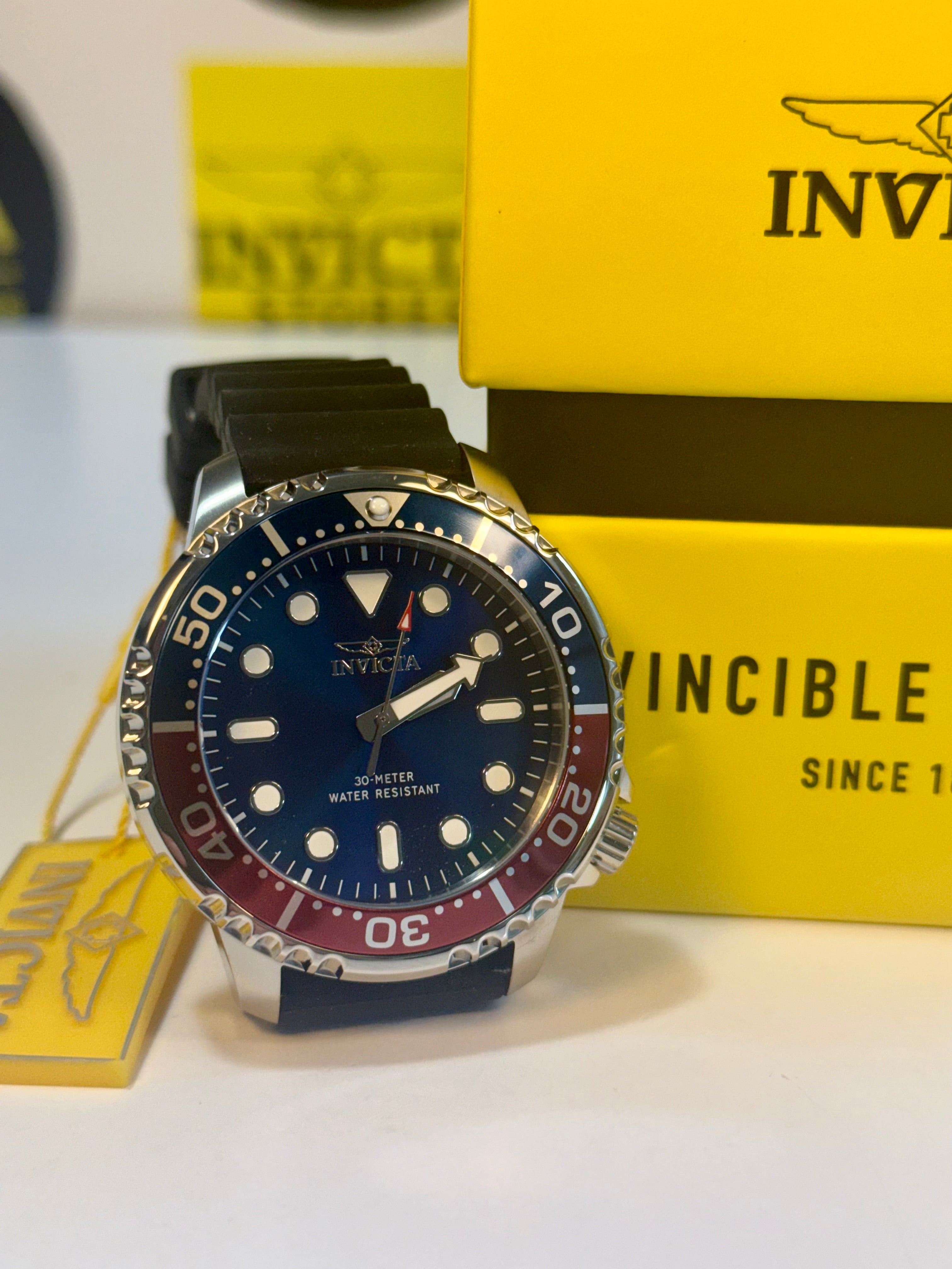 Invicta Mens Pro Diver 48mm Silicone band watch with blue dial and black strap on a white surface with yellow Invicta packaging in the background.
