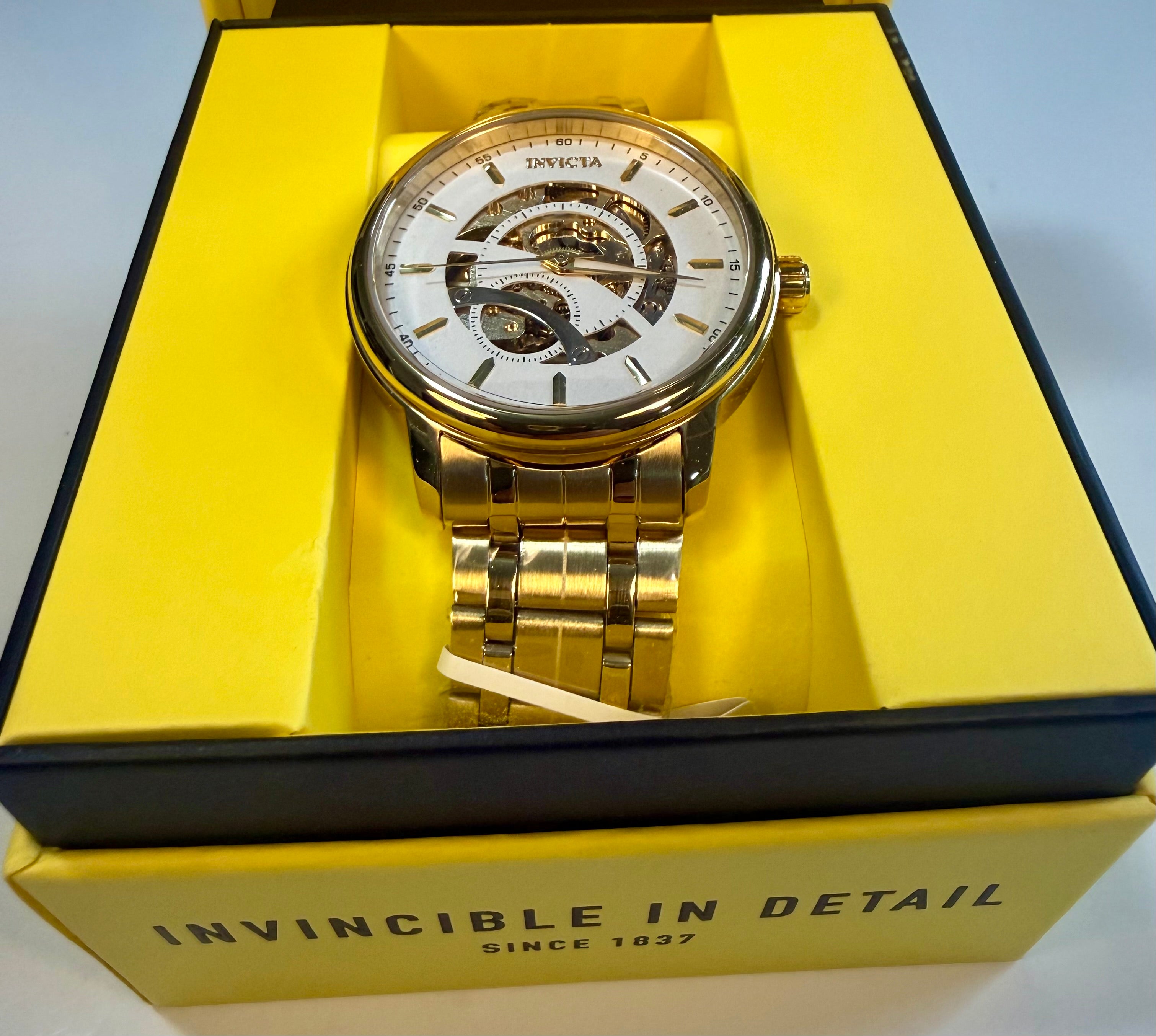 Invicta Mens Automatic Objet' De Art  Gold wristwatch with white face in a yellow box labeled 'Invincible in Detail'.