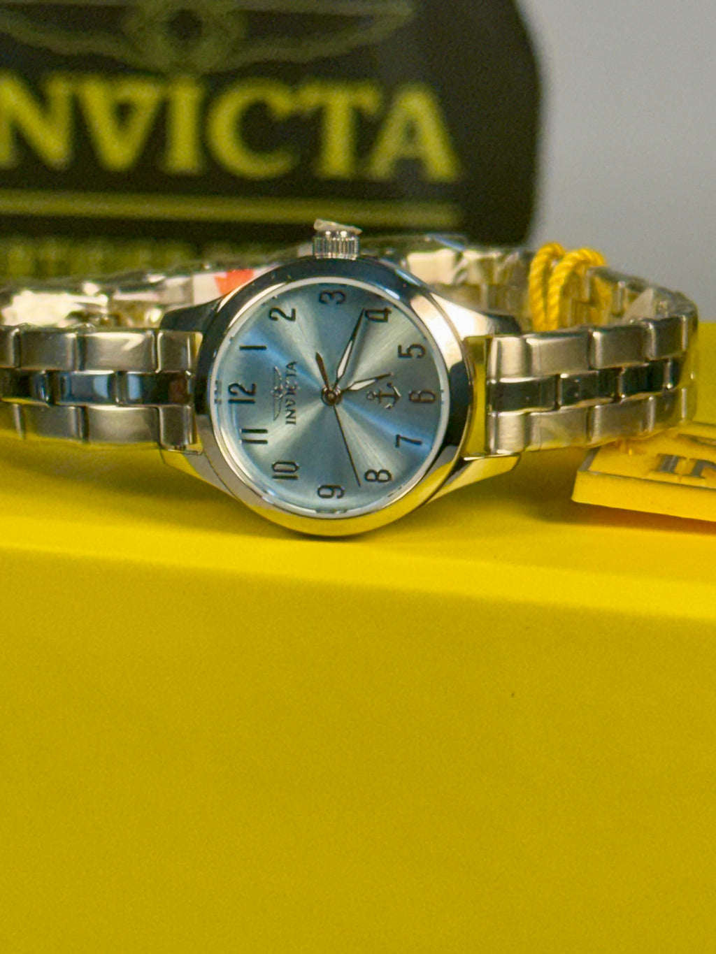 Invicta Ladies Ocean Voyage Invicta watch on a yellow surface with a blurred Invicta logo in the background