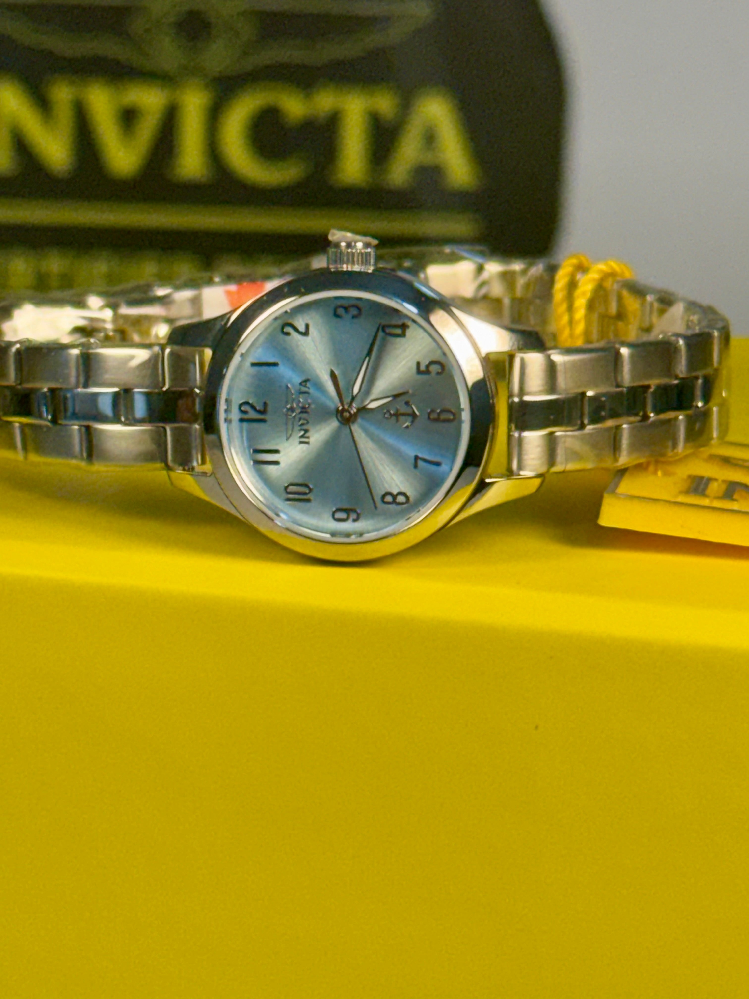 Invicta Ladies Ocean Voyage Invicta watch on a yellow surface with a blurred Invicta logo in the background